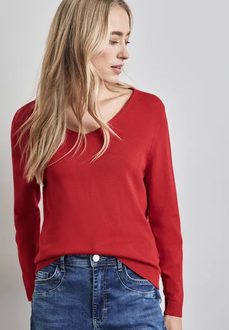 Frau mit langem blondem Haar, rotem Pullover und blauer Jeans steht vor grauem Hintergrund.